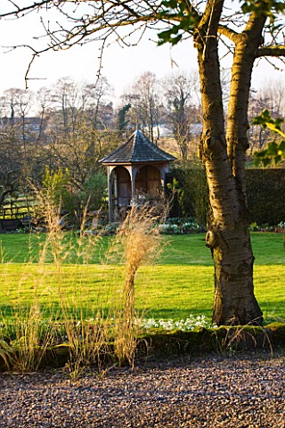 DIAL_PARK__WORCESTERSHIRE_VIEW_ACROSS_GRAVEL_DRIVE_AND_LAWN_TO_THE_GAZEBO_WITH_ORYZOPSIS_MILEEACEA_I