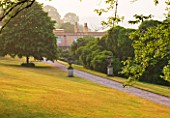 KILLERTON  DEVON: THE NATIONAL TRUST - PATH THROUGH LAWNS AND TREES IN THE GARDEN WITH THE WEST FRONT OF THE HOUSE - MORNING LIGHT