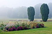 GLYNDEBOURNE, EAST SUSSEX: THE MAIN LAWN WITH BENCHES AND YEWS, BORDER WITH SEDUMS AND GAURA LINDHEIMERI - SHEEP BEYOND IN FIELD, COUNTRY GARDEN