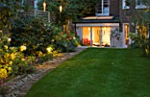 NOTTING HILL HOUSE, LONDON.GARDEN DESIGN BY BUTTER WAKEFIELD. VIEW OF REAR OF HOUSE WITH LAWN AND GARDEN LIGHTING IN BORDER.ILLUMINATE,LIT,UPLIGHTING,NIGHT,EVENING,GLOW,ATMOSPHERE