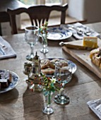 COMMON FARM FLOWERS. SOMERSET: FESTIVE TEA TABLE SET WITH INDIVIDUAL PLACE SETTINGS OF BRITISH FLOWERS IN SHERRY GLASSES ; SPRIGS OF HOLLY  ROSEHIPS AND SCENTED TAZETTA NARCISSI