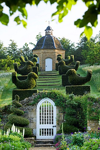 ROCKCLIFFE_HOUSE_GLOUCESTERSHIRE_VIEW_THROUGH_WHITE_GATE_TO_ROW_OF_CLIPPED_TOPIARY_BIRDS_LEADING_UP_