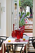 BEN DE LISI HOUSE AND GARDEN  LONDON: VIEW OUT OF KITCHEN WITH TABLE THROUGH THE BACK DOOR TO GARDEN WITH TABLE AND COLLECTION OF RED GLASS AND VINTAGE MURANO PIECES