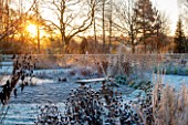 ELLICAR GARDENS, NOTTINGHAMSHIRE: GRASSES AND WOODEN BENCH WITH VIEW ACROSS THE POOL AT SUNRISE, WINTER, FROST