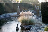 SEDGWICK PARK, WEST SUSSEX. COLUMNS OF YEW BESIDE THE LONG CANAL ALSO KNOWN AS THE WHITE SEA WITH PAMPAS GRASS. WINTER, GARDEN, FROST, JANUARY, WATER