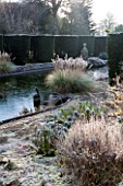 SEDGWICK PARK, WEST SUSSEX. COLUMNS OF YEW BESIDE THE LONG CANAL ALSO KNOWN AS THE WHITE SEA WITH PAMPAS GRASS AND FROSTED EUPHORBIAS. WINTER, GARDEN, FROST, JANUARY, WATER