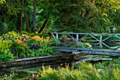 ABLINGTON MANOR  GLOUCESTERSHIRE: WOODN FOOTBRIDGE OVER THE RIVER COLN WITH ISLAND BED OF HOT PERENNIALS  RIVER  WATER  CLASSIC COUNTRY GARDEN  FLOWERS