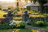 PETTIFERS GARDEN, OXFORDSHIRE: EARLY MORNING VIEW FROM THE BACK OF THE HOUSE WITH LAWN, GRAVEL PATH AND LEAD URNS TO THE COUNTRYSIDE BEYOND