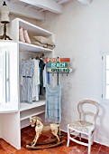 CIUTADELLA MENORCA, SPAIN: EVELYNE MANDEL HOUSE - BEDROOM WITH WHITE PAINTED WALLS - BUILT IN WARDROBE WITH DRESSES AND PILLOWS, ANTIQUE ROCKING HORSE, WHITE CHAIR