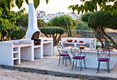 JONATHAN BAILLIE GARDEN, ALAIOR, MENORCA: BUILT IN BARBEQUE AND TABLE WITH METAL CHAIRS, SURROUNDED BY CLIPPED OLIVE TREES. FOOD, DINING, ENTERTAINING AREA, OUTDOOR LIFESTYLE