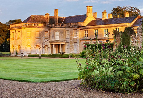 LAMPORT_HALL_NORTHAMPTONSHIRE_THE_HALL_SEEN_ACROSS_THE_LAWN_WITH_HOLLYHOCKS_IN_THE_FOREGROUND__FLOWE