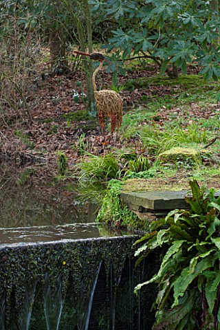RODE_HALL_AND_GARDENS_CHESHIRE_STEW_POND_IN_WOODLAND_WITH_WILLOW_HERON_SCULPTURE_COUNTRY_GARDEN_FEBR
