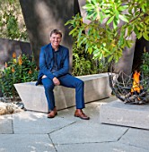 CHELSEA FLOWER SHOW 2016: TELEGRAPH GAREDEN DESIGNED BY ANDY STURGEON - ANDY STURGEON SITTING ON LIMESTONE SEAT / BENCH ON LIMESTONE PATIO, TERRACE WITH FIREPIT