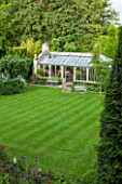 THE LODGE, BURFORD, OXFORDSHIRE: THE PELARGONIUM FILLED CONSERVATORY WITH BETULA PENDULA ON LAWN AND EVERGREEN TREES. WITH METAL TABLE & CHAIRS ON STRIPED LAWN. BEAUTIFUL GARDEN