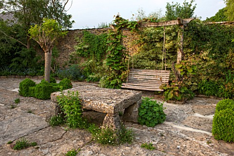 OXLEAZE_FARM_OXFORDSHIRE_SWING_SEAT_AND_STONE_TABLE_WITH_ROBINIA_INERMIS_AND_VITIS_COGNETIAE_COMING_