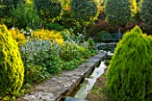 EASTLEACH HOUSE, GLOUCESTERSHIRE: VIEW DOWN THE RILL TO TREES - SORBUS ARIA LUTESCENS. WATER, CANAL, PLATYCLADUS ORIENTALIS AUREA NANA, EVENING LIGHT, COUNTRY GARDEN