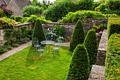 CALENDARS:BURFORD,OXFORDSHIRE:CLIPPED YEW CONES ON LAWN. WALL WITH TABLE & CHAIRS.BORDER WITH ERIGERON KARVINSKIANUS,GERANIUM ROZANNE,SHASTA DAISIES & POTS OF FERNS ON STEPS.SUMMER
