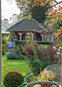 THE COACH HOUSE, SURREY: REAR/BACK GARDEN IN AUTUMN DESIGNED BY BARBARA BROOKS. GARDEN BUILDING/POTTING SHED WITH FURNITURE. SHRUBS, GRASSES, PERENNIALS, LAWN, BOX BALL.