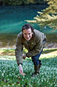 COLESBOURNE PARK, GLOUCESTERSHIRE: HEAD GARDENER ARTHUR COLE BY THE LAKE WITH SNOWDROPS