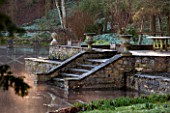 ABLINGTON MANOR, GLOUCESTERSHIRE: VIEW ACROSS COLN RIVER TO STONE STEPS AND CONTAINERS. CLASSIC, COUNTRY GARDEN, COTSWOLDS, ROMANTIC, ROMANCE, WINTER, EARLY SPRING