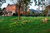MORTON HALL GARDENS, WORCESTERSHIRE: THE HALL SEEN FROM THE MONONPTEROS. PARKLAND MEADOW, DAFFODILS, NARCISSUS, COLONNADE, PARK, SUNRISE, SPRING, BENCH, WOODEN, SEAT