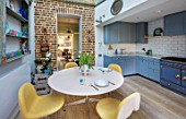 LONDON HOUSE DESIGNED BY JULIE SIMONSEN. AZURE BLUE KITCHEN BY LAURENCE PIDGEON WITH LA CORNUE RANGE OVEN ADJACENT TO MORNING ROOM WITH EXPOSED BRICKWORK AND CIRCULAR TABLE AND YELLOW CHAIRS.