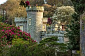 CAERHAYS CASTLE, CORNWALL: THE CASTLE SEEN FROM THE GARDEN FRAMED BY RHODODENDRON CORNISH RED. ENGLISH, COUNTRY, GARDEN, SPRING, APRIL