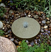 SIMPLE MILLSTONE FOUNTAIN SURROUNDED BY PEBBLES IN DALES STONE COMPANY GARDEN. CHELSEA 1994