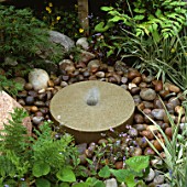 SIMPLE MILLSTONE FOUNTAIN SURROUNDED BY PEBBLES IN DALES STONE COMPANY GARDEN. CHELSEA 1994.