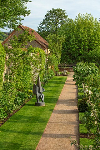 CHILWORTH_MANOR_SURREY_THE_WALLED_GARDEN_GRAVEL_PATH_PAST_LAWN_AND_SCULPTURE_IN_SPRING