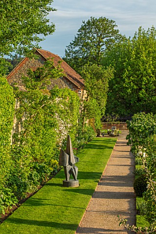 CHILWORTH_MANOR_SURREY_THE_WALLED_GARDEN_GRAVEL_PATH_PAST_LAWN_AND_SCULPTURE_IN_SPRING