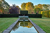 RADCOT HOUSE, OXFORDSHIRE: THE LONG POND. YEW HEDGES, HEDGING, POOL, CANAL, AUTUMN, REFLECTIONS, REFLECTED, STONE, URN, WHITE METAL SEAT, SEATING