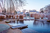 THE OLD RECTORY, QUINTON, NORTHAMPTONSHIRE: DESIGNER ANOUSHKA FEILER: VIEW ACROSS LAKE TO RECTORY WITH GRASSES AND WOODEN PONTOON, FROST, WINTER, FROSTY GARDEN, ENGLISH, COUNTRY