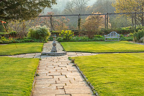 GRAVETYE_MANOR_SUSSEX_SPRING_APRIL_COUNTRY_GARDEN_LUTYENS_BENCH_SEAT_ON_LAWN_PATHS_AND_STONE_SUNDIAL
