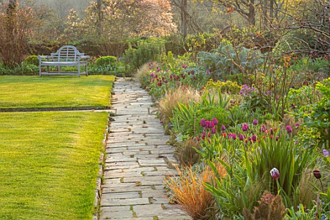 GRAVETYE_MANOR_SUSSEX_SPRING_APRIL_COUNTRY_GARDEN_LUTYENS_BENCH_SEAT_ON_LAWN_PATHS_AND_BORDER_FILLED