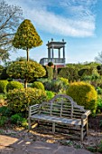 THE LASKETT GARDENS, HEREFORDSHIRE. DESIGNER ROY STRONG - THE SERPENTINE WALK, WOODEN BECNCH, SEAT, CLIPPED TOPIARY HOLLIES, YEW, BELVEDERE, BUILDINGS, SPRING, APRIL