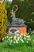 THE LASKETT GARDENS, HEREFORDSHIRE. DESIGNER ROY STRONG - CHILSTONE STAG ON PEDESTAL WITH DAFFODILS IN THE CHRISTMAS ORCHARD, APRIL, SPRING